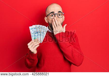 Young bald man holding peruvian sol banknotes covering mouth with hand, shocked and afraid for mistake. surprised expression 