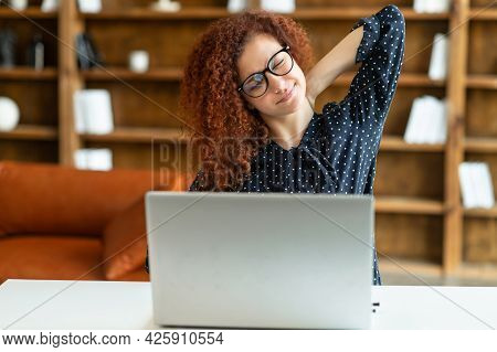 Carefree Attractive Red-haired Curly Woman In Eyeglasses Stretching Arms Sitting At Desk In The Offi