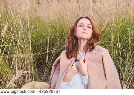 An Adult Woman In A Beige Coat Sits On The Autumn Grass In A Field