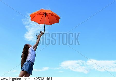 Beautiful Young Girl White T-shirt And Denim Shorts Throws Up An Orange Umbrella. A Flying Umbrella 