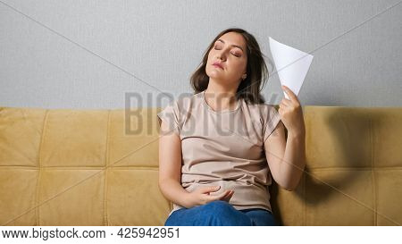 Young Woman Suffering From Heat Fan Herself With A Sheet Of Paper While Sitting On The Couch.