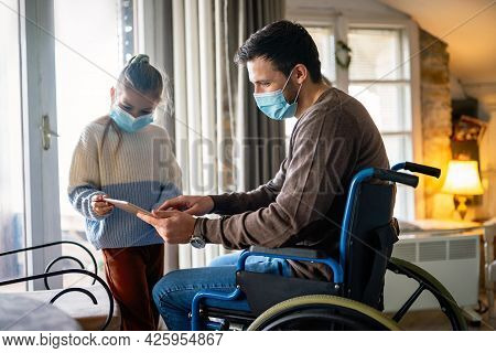 Father With Disability In Wheelchair Using Tablet At Home With Child While Wearing Masks.