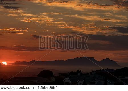 The Sun Is Setting Behind Fish Hoek Accross False Bay. The Back Of Table Mountain And Devils Peak Ar