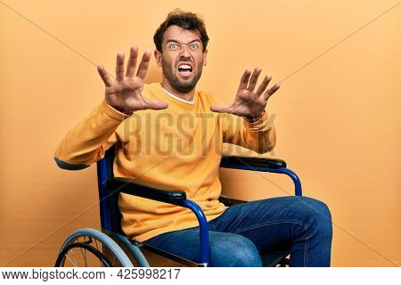 Handsome man with beard sitting on wheelchair afraid and terrified with fear expression stop gesture with hands, shouting in shock. panic concept. 