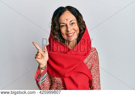 Middle age hispanic woman wearing tradition sherwani saree clothes with a big smile on face, pointing with hand and finger to the side looking at the camera. 
