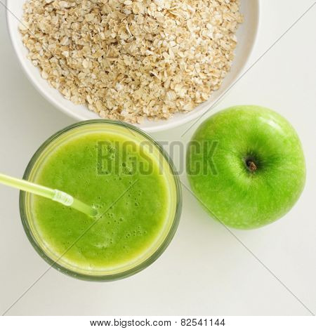 high-angle shot of an apple, a glass with a green smoothie and a bowl with oatmeal cereal on a white table