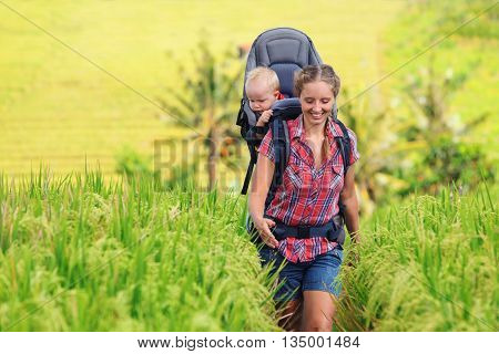 Nature walk in green rice terrace field. Happy mother hold little traveller in carrying backpack. Baby ride on woman back. Travel adventure hiking with child carrier family summer vacation on Bali.