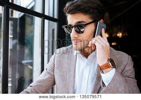 Seious young businessman in sunglasses talking on mobile phone in cafe