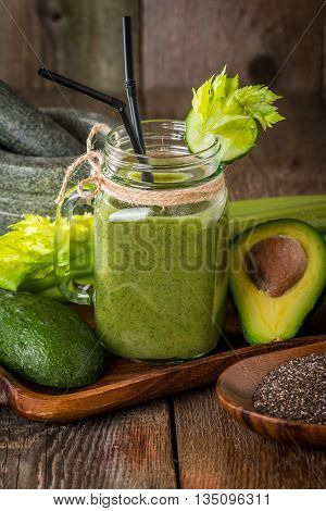 Healthy green juice smoothie with straw surrounded by avocado, cucumber,  celery and chia seeds on wooden background