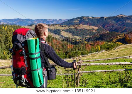 Hiker with Backpack and Walking Pole Standing on Grassy Meadow Observing Mountains Scenery of Autumnal Colorful Forest