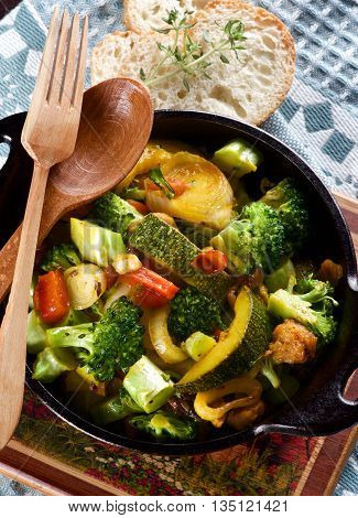 Tasty Colorful Vegetables Ragout with Zucchini Carrots Broccoli Leek and Red Bell Pepper in Black Iron Stewpot with Wooden Spoon and Fork and Bread with Thyme closeup on Blue Napkin