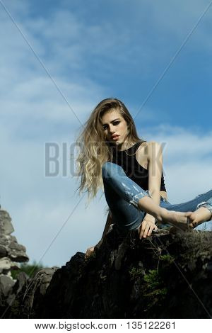 Beautiful girl with slim body and long hair sitting on stone over blue sky with clouds