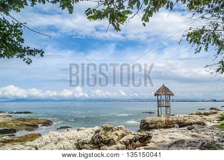 Expansive ocean view from rocky beach with palapa lifeguard shack and blue sky with puffy white clouds.