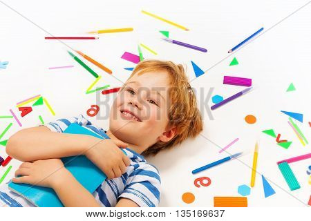 Close up picture of smiling five years old boy with book, having a rest among heap of stationery