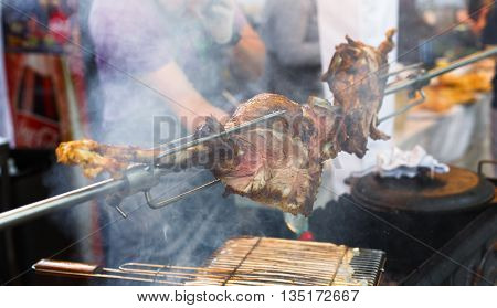 Meat barbecue picnic at country fair. Lamb leg roasted at spit. Lamb grill, big piece of meat at rolling skewer. Street vendor, professional Chef cooking food bbq, unrecognizable. Cooking, catering