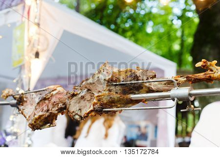Meat barbecue picnic at country fair. Lamb leg roasted at spit. Lamb grill, big piece of meat at rolling skewer. Fresh cooked food bbq for sale.