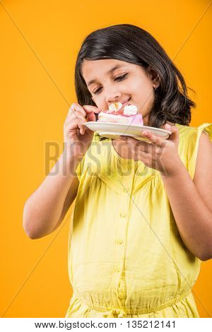 portrait of Indian kid eating cake or pastry, cute little girl eating cake, girl eating strawberry cake or pastry over yellow background