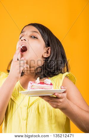 portrait of Indian kid eating cake or pastry, cute little girl eating cake, girl eating strawberry cake or pastry over yellow background