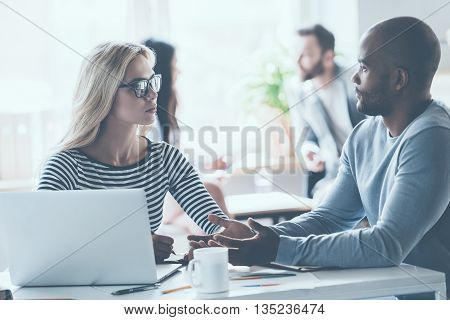 Discussing some business issues. Two young business people discussing something while sitting at the office desk together while their colleagues sitting in the background
