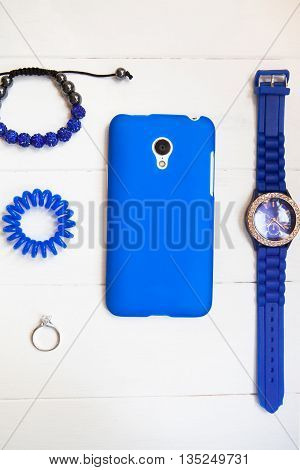 Smartphone in blue case with ring braslets with rhinestones scrunchy blue watch on white wooden background with copy space. Top view flat lay