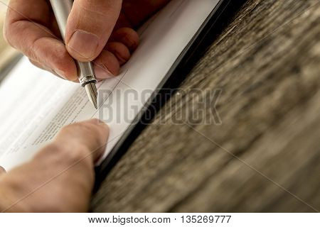 Low angle closeup view of male hand signing contract document or application form with a pen on textured wooden desk.
