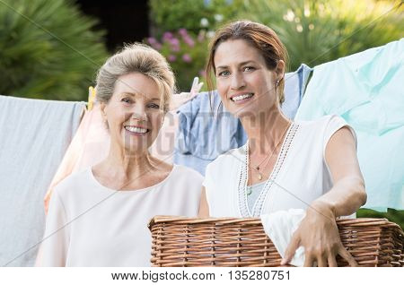 Daughter holding laundry basket standing in front of the laundry hanging. Old mother and daughter looking at camera with launfry on background. Happy women enjoying house work.