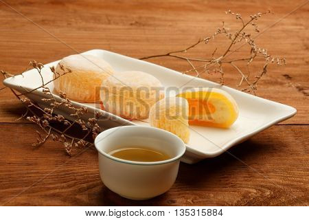 Beautiful mango mochi rice cakes on white square ceramic plate with green tea in chinese porcelain cup standing on brown wooden table decorated with dried flowers.