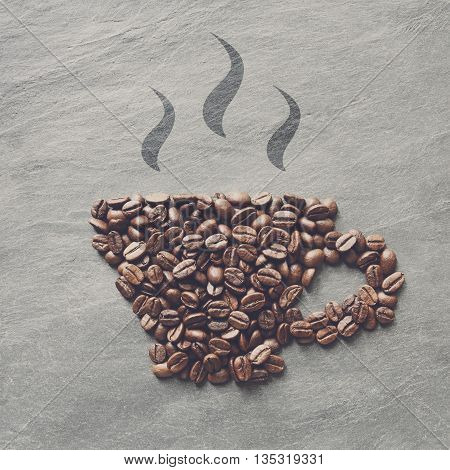 Cup from coffee beans with smoke. Heap of roasted coffee bean on grey stone surface texture. Coffee shop or cafe background. Natural stone and cup from beans. Soft color toning, top view