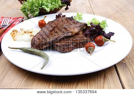 meat food : two grilled steak on green lettuce salad , with roast onion and red hot chili peppers , on dish over wooden table