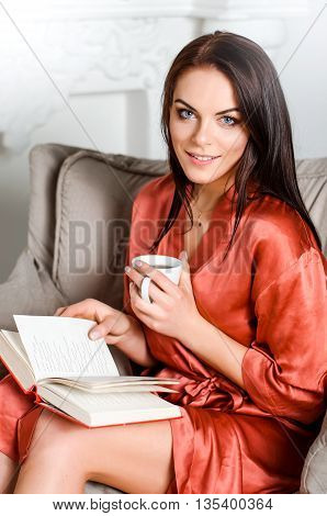 young woman in the brown-haired sholkom dressing gown sitting on a chair in his house reading a book and drinking tea or coffee