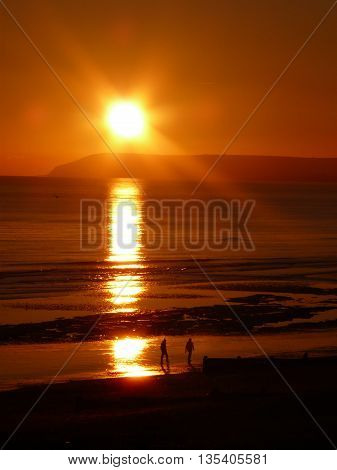 A golden sun setting over a beach where two people are walking