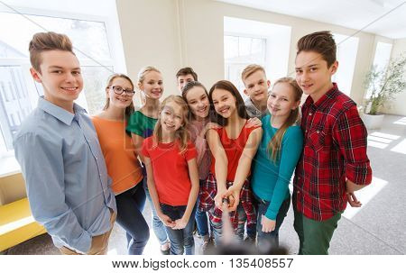 education, school, technology and people concept - group of happy smiling students taking picture with smartphone selfie stick in corridor