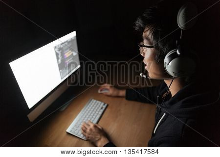 Concentrated asian male gamer sitting and playing computer game in dark room
