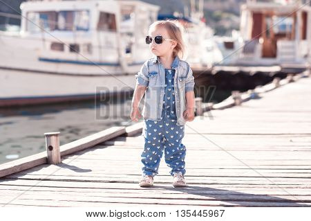 Stylish baby girl wearing denim suit and jacket at wooden pier. Summer season. Child standing outdoors.