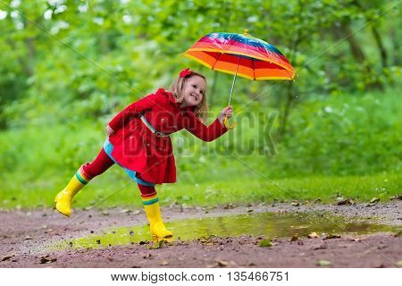 Little girl playing in rainy summer park. Child with colorful rainbow umbrella waterproof coat and boots jumping in puddle in the rain. Kid walking in autumn shower. Outdoor fun by any weather