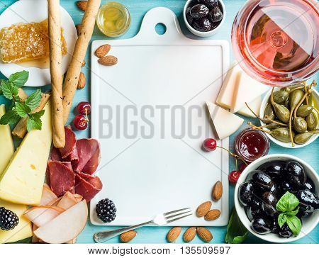Summer wine snack set. Glass of rose, meat, cheese, olives, honey, bread sticks, nuts, capers and berries with white ceramic board in center, blue wooden background, top view, copy space