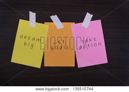 Word quotes of Dream Big Set Goals Take Action on sticky color papers against wooden textured background.