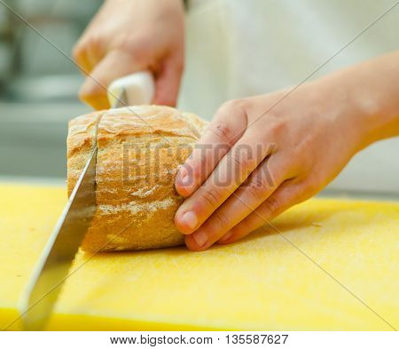 Cutting slices from loaf of bread sitting on yellow surface, hand holding large knife.