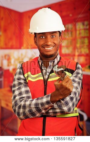 Young engineer carpenter wearing helmet, square pattern flanel shirt with red safety vest, holding small handheld electric saw smiling to camera.