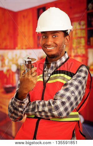 Young engineer carpenter wearing helmet, square pattern flanel shirt with red safety vest, holding small handheld electric saw smiling to camera.
