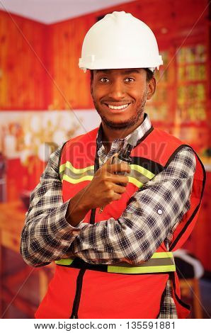 Young engineer carpenter wearing helmet, square pattern flanel shirt with red safety vest, holding small handheld electric polisher tool smiling to camera.