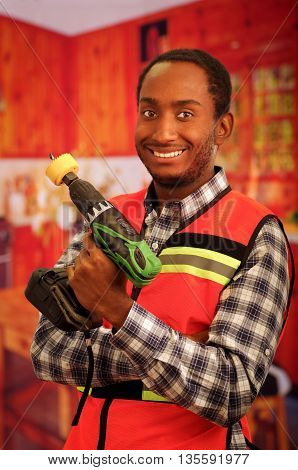 Young engineer carpenter wearing square pattern flanel shirt with red safety vest, holding handheld electric power drill smiling to camera.