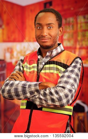 Young carpenter worker wearing square pattern flanel shirt and red safety vest, posing with arms crossed, smiling to camera.