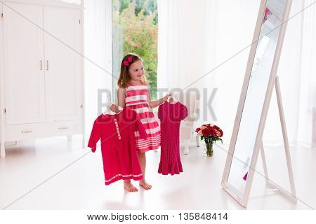 Little girl choosing dresses in white bedroom. Child watching mirror reflection holding pink dress choosing outfit. Girls nursery. Shopping clothing for kids. Dressing room interior for children.