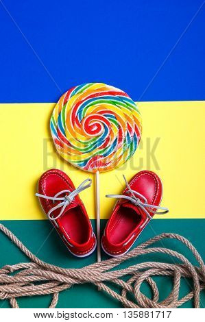 Small red boat shoes near big multi-colored lollipop and rope on colored background. Top view. Close up.