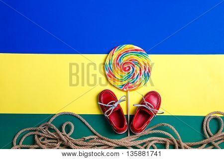 Small red boat shoes near big multi-colored lollipop and rope on colored background. Top view. Copy space frame