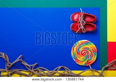 Small red boat shoes near big multi-colored lollipop and rope on colored background. Top view. Copy space frame