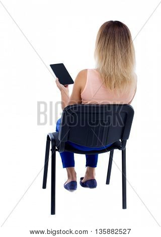 back view of woman sitting on chair and looks at the screen of the tablet. Isolated over white background. Blonde in blue pants sitting on an office chair holding a tablet computer.