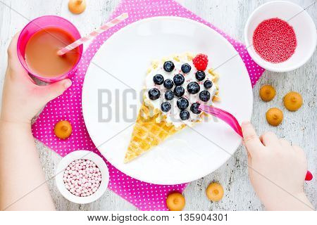 Creative baby breakfast idea - wafer with mascarpone cream and berries in shape ice cream. Child eating