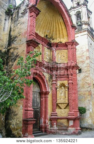 Ancient arched church entrance of gold and rust painted stone with wooden door and statuary next to white stone bell tower.